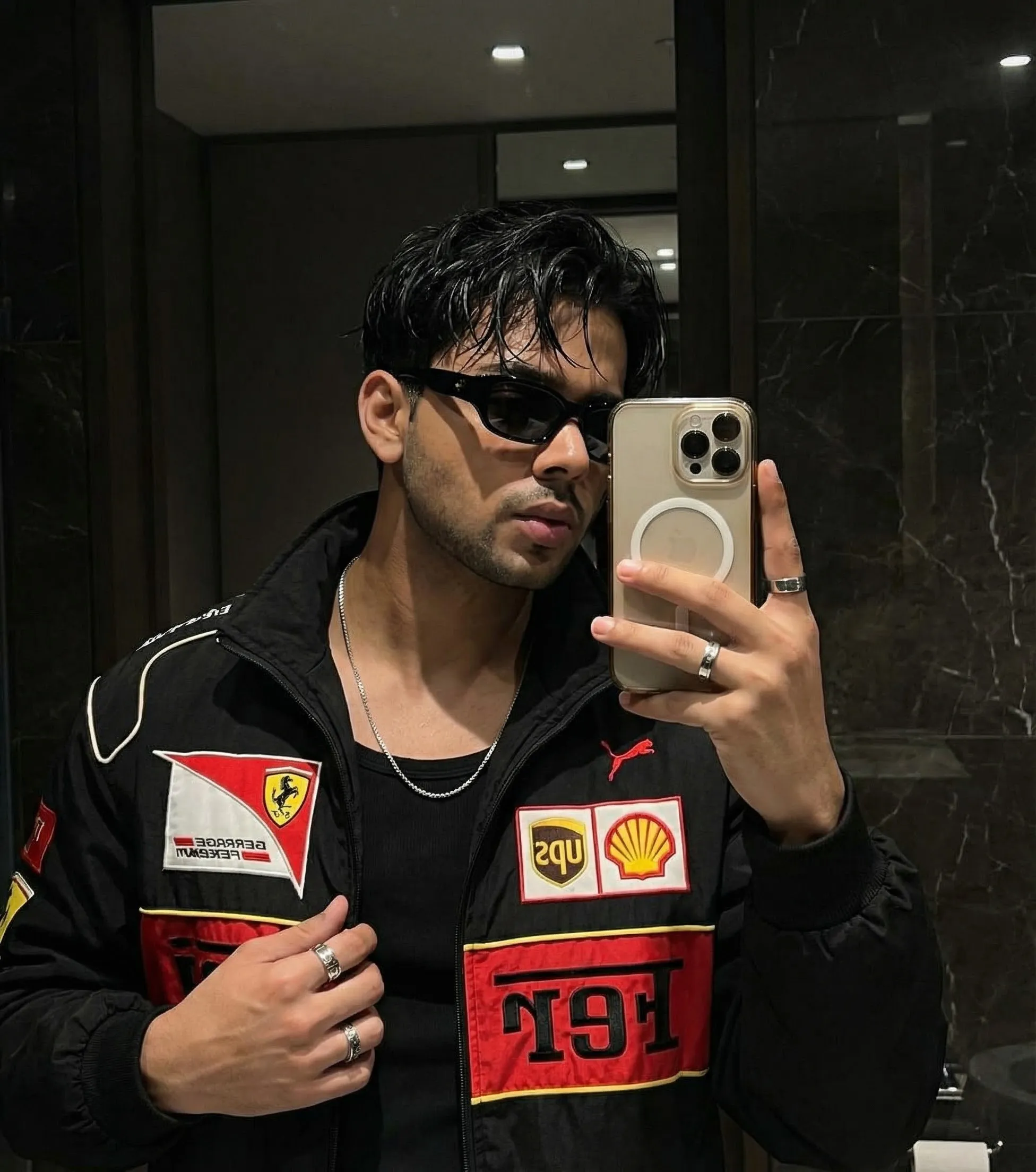 Mirror selfie photo editing prompt for boys - young man in a black Ferrari racing jacket with red and yellow patches, black sunglasses and silver rings taking a phone selfie in a dark marble bathroom